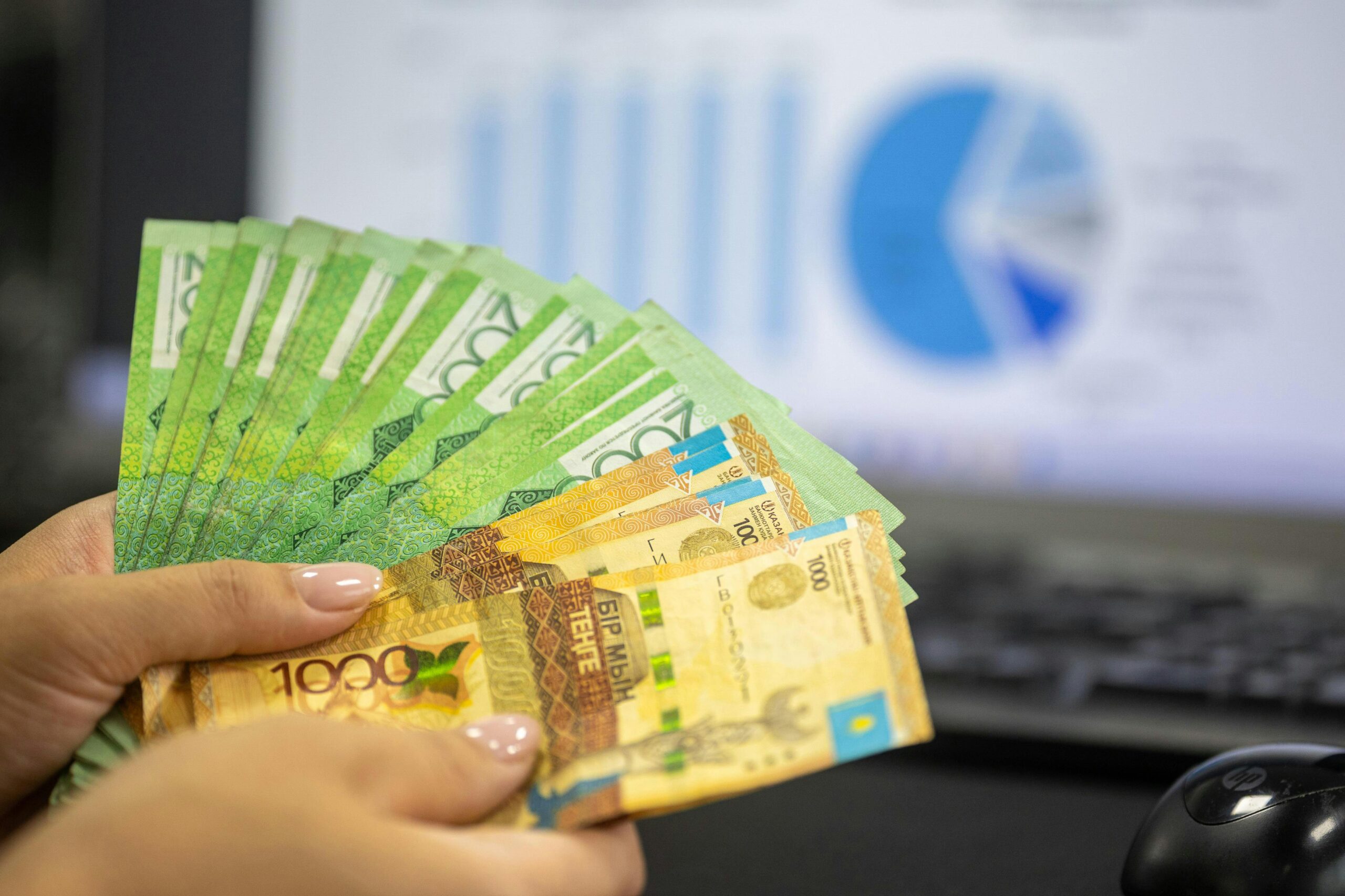 Nahaufnahme von Händen, die kasachische Tenge-Scheine halten, mit Finanzdiagrammen im Hintergrund.